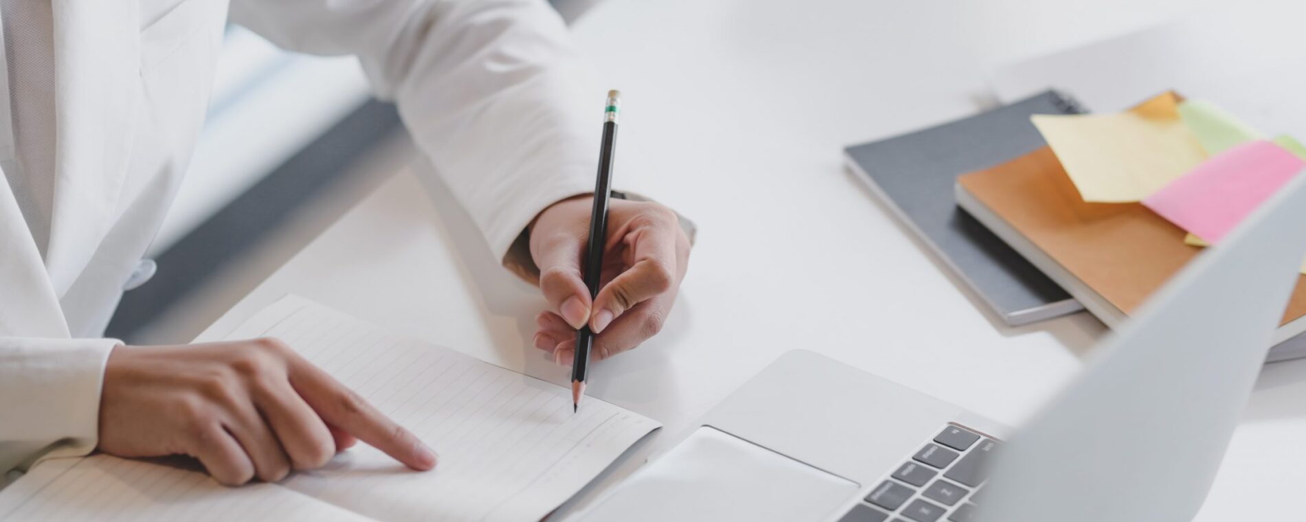 Cropped shot of businesswoman writing on notebook while working on laptop with office supplies on white table